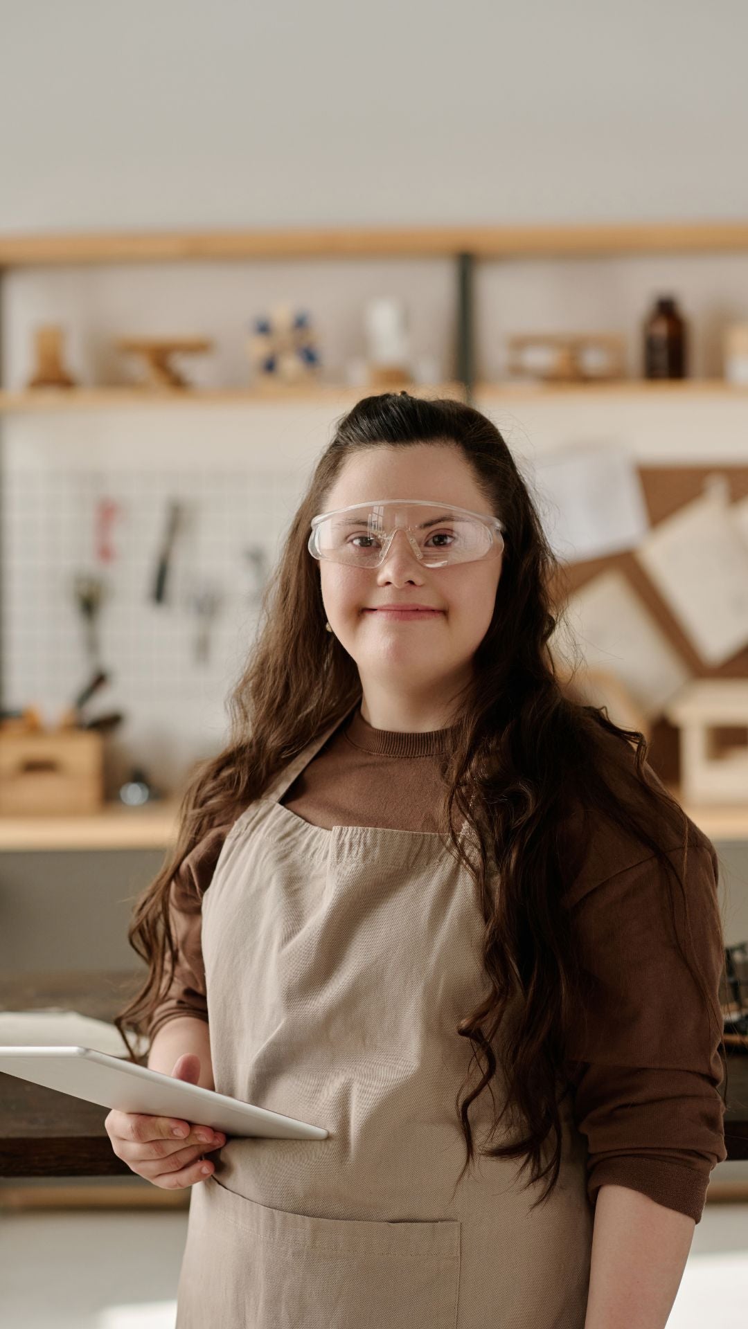 Vertical image of girl with down syndrome smiling at camera while using digital tablet in workshop